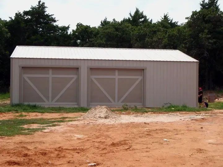 Two-bay metal garage with white doors and roof on a dirt lot, trees in the background. A person stands to the right.