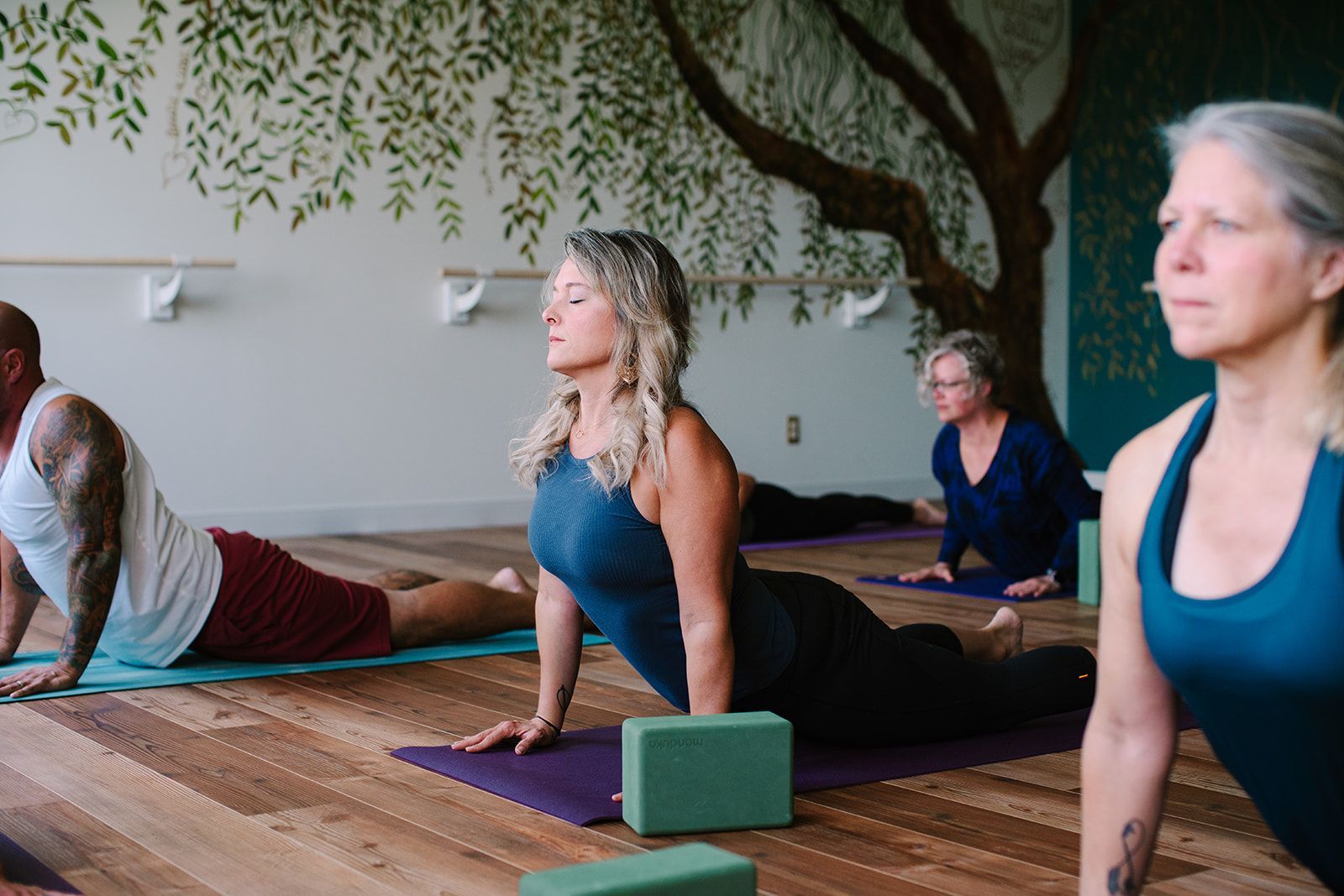 People doing yoga poses on mats in a studio with wood floors and a painted tree mural.
