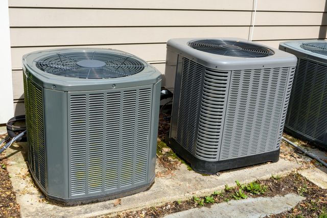 Three outdoor air conditioning units beside a house wall on a concrete pad with weeds nearby