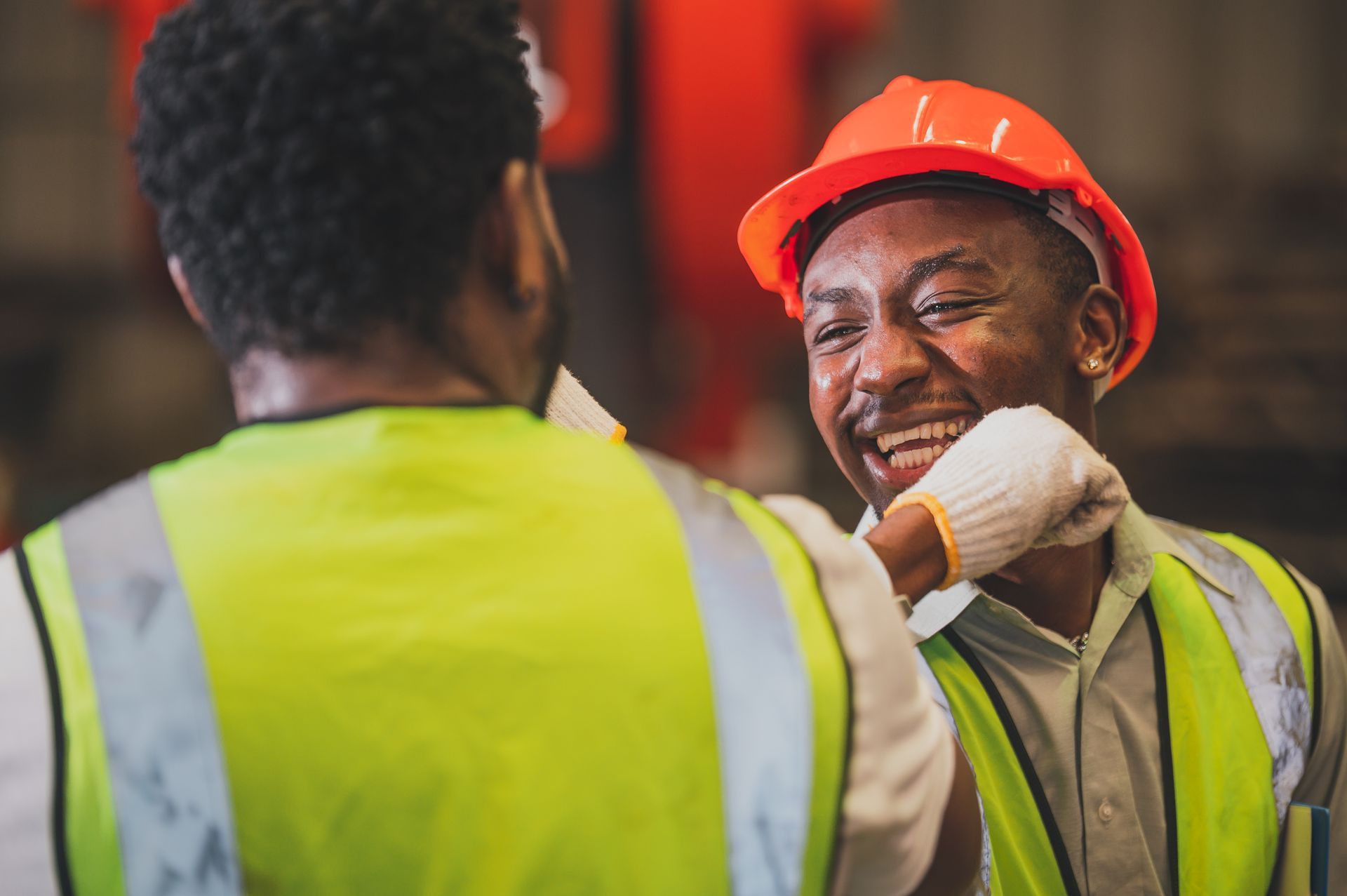 Two construction workers in safety vests and hard hats smile during a site conversation