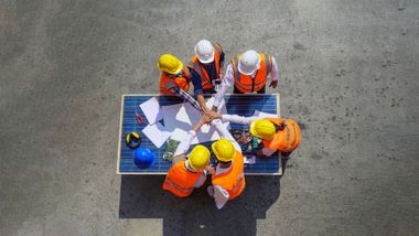 Workers in orange vests and yellow hard hats reviewing plans around a blue table on concrete.