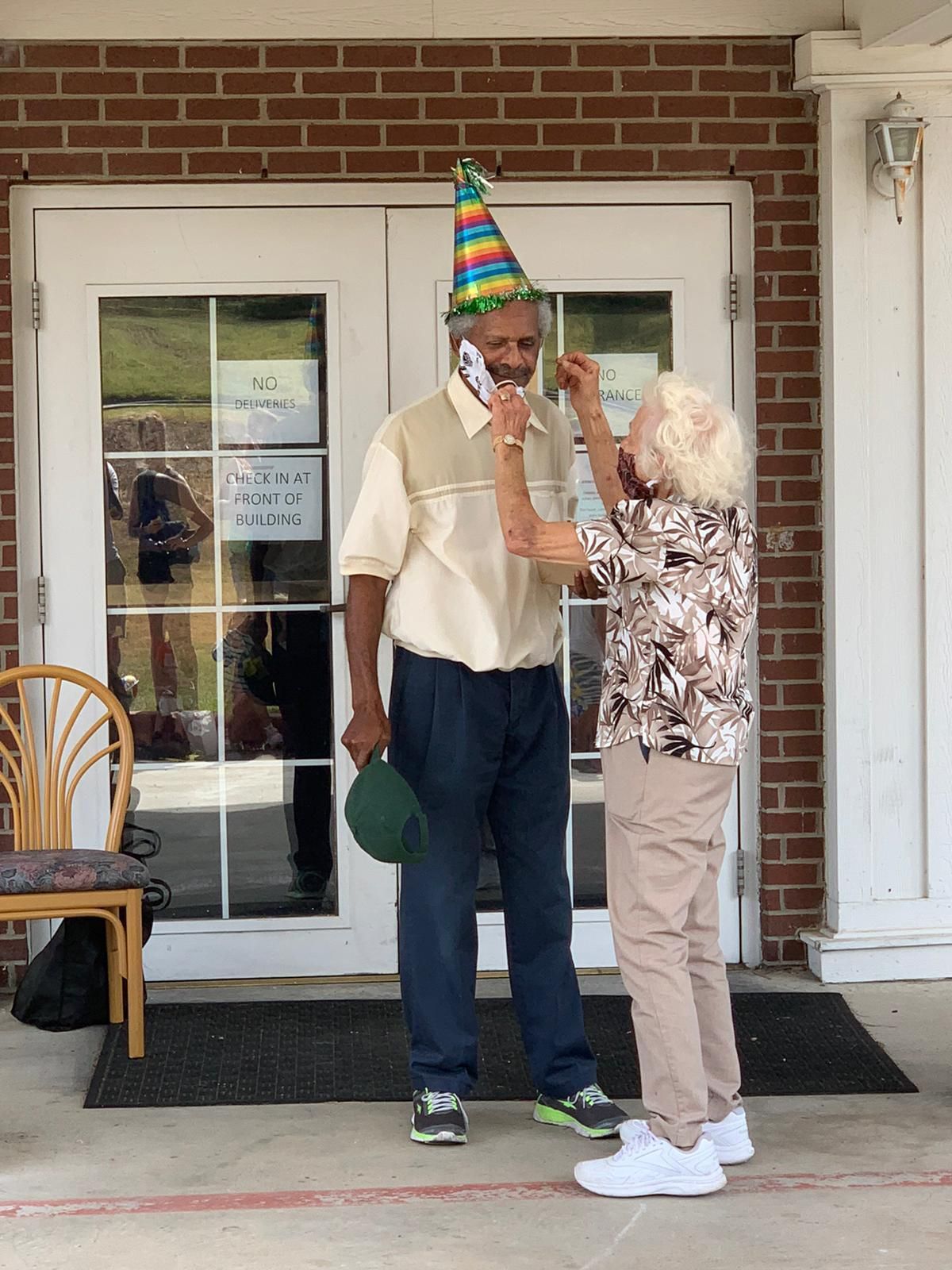 A person adjusts a party hat on another person's head outside a building with brick facade.