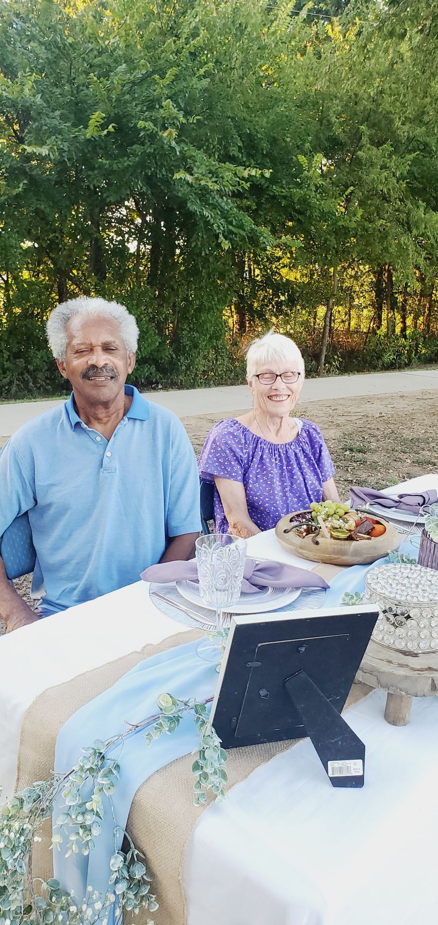 Two people sitting at a table set for a meal. Trees in the background.