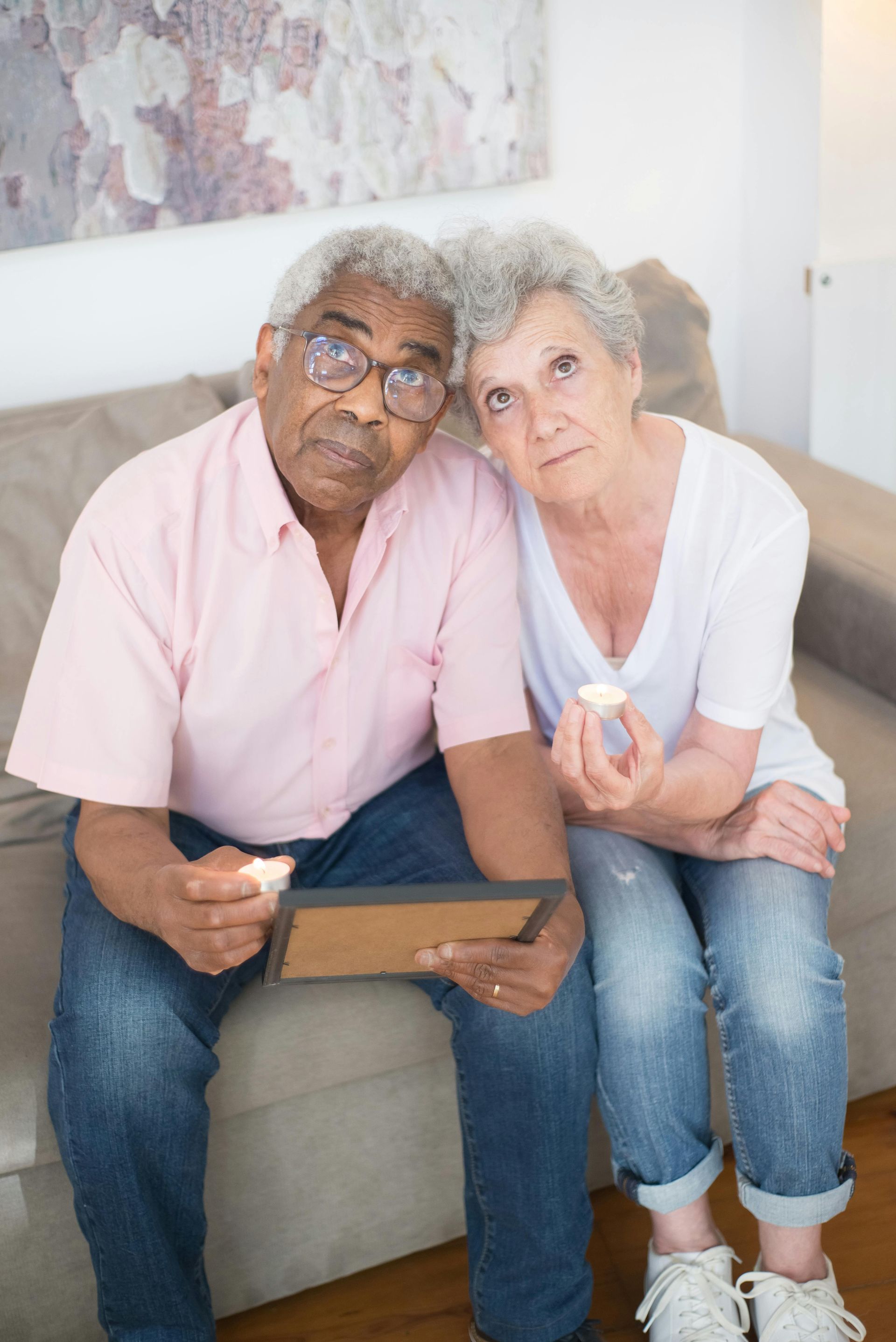 Elderly couple on couch, man holds photo, woman holds a pill, both look concerned.