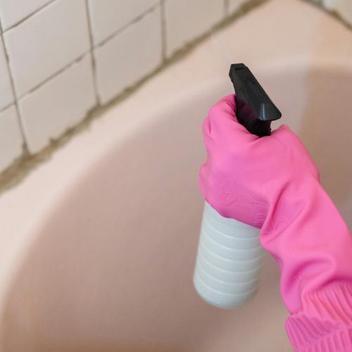 A person wearing pink gloves is holding a spray bottle in front of a bathtub.