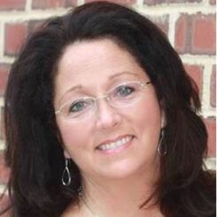 A woman wearing glasses and earrings is smiling in front of a brick wall.