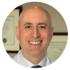 A bald man in a white coat and tie smiles for the camera