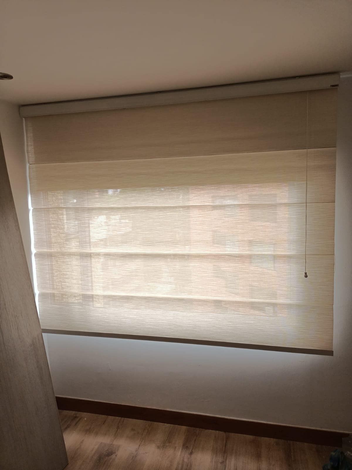 Estores romanos color beige en una ventana de una habitación con paredes blancas y molduras de madera.