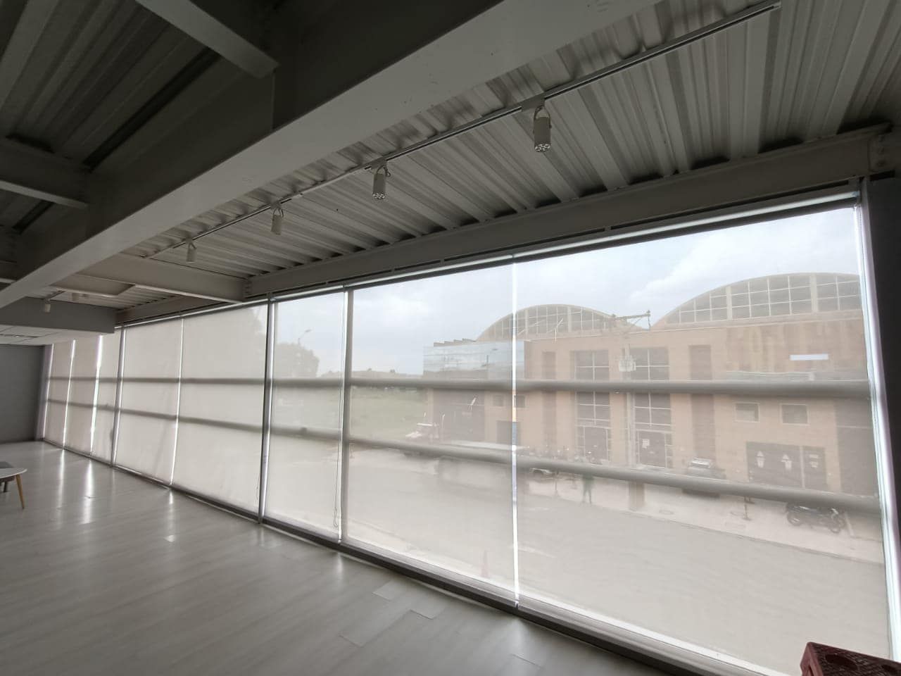 Vista interior de una habitación con grandes ventanas, parcialmente cubiertas por persianas enrollables color beige, con vistas a un edificio.