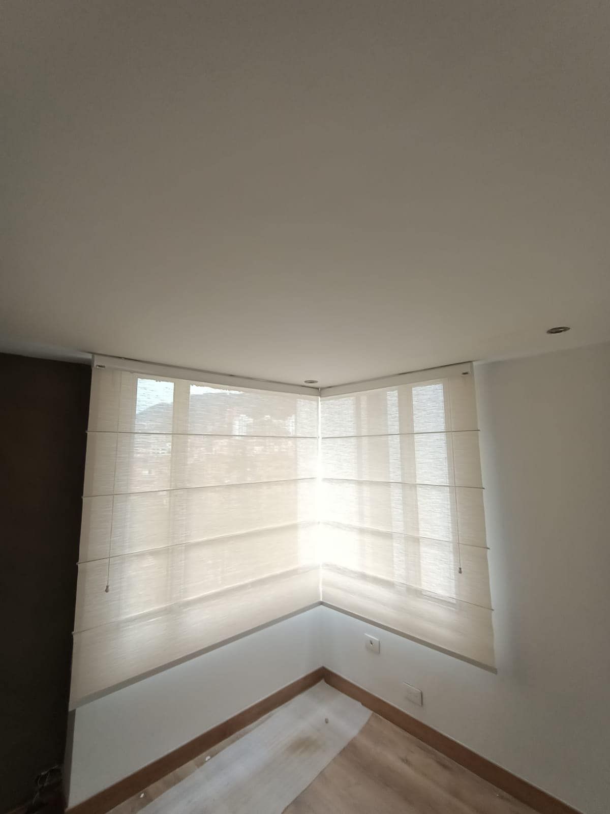 Ventana esquinera con estores romanos claros. La ventana ofrece vistas a lo lejos; la habitación tiene paredes claras y suelo de madera.