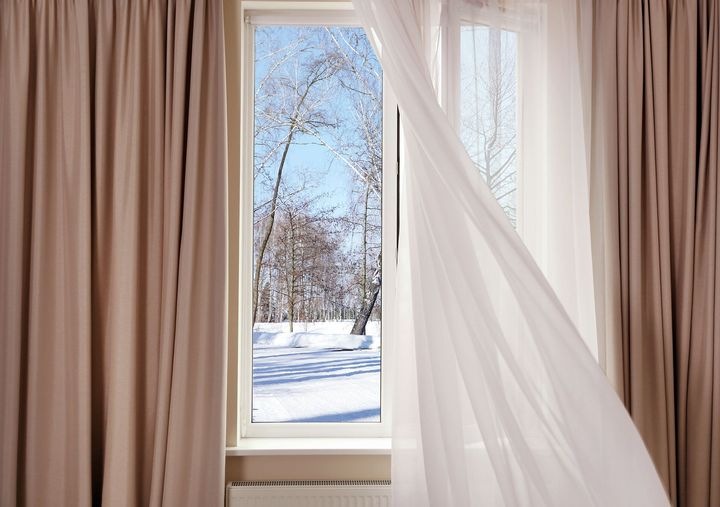 Ventana con una cortina blanca transparente que ondea al viento, dejando ver árboles cubiertos de nieve y un cielo azul; unas cortinas color beige enmarcan la ventana.
