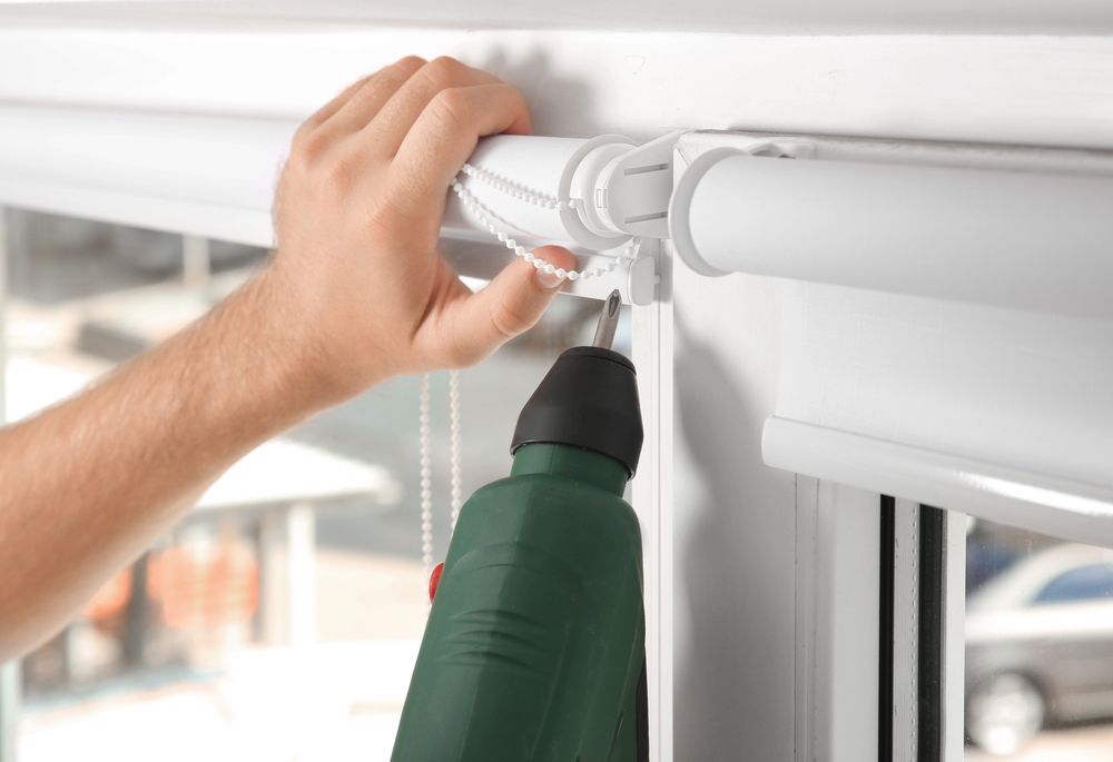 Persona utilizando un taladro para instalar una persiana enrollable blanca en el marco de una ventana.