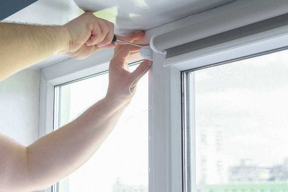 Persona utilizando un destornillador para instalar una persiana blanca, cerca de un marco de ventana blanco.