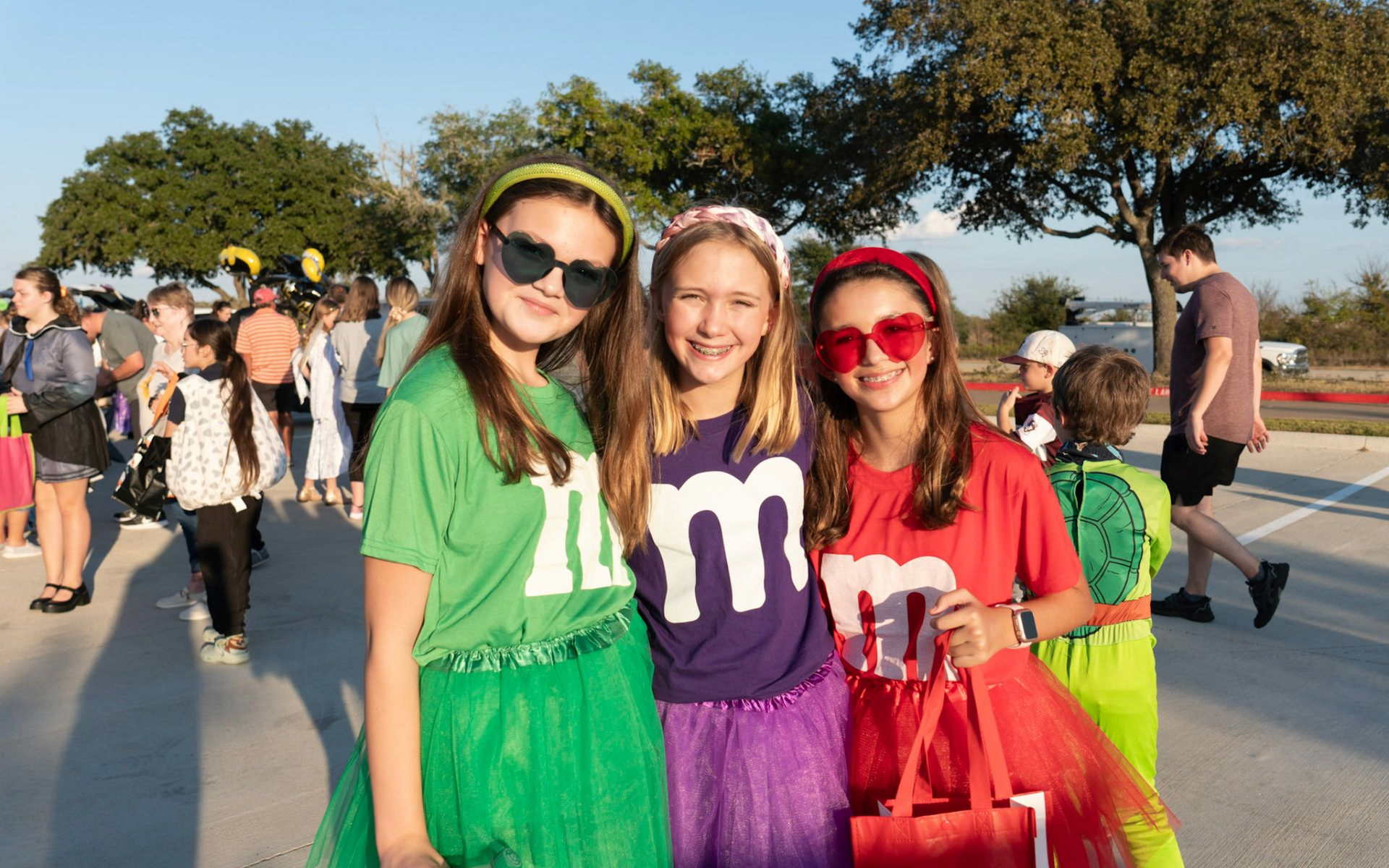 Three girls in M&M costumes smile outdoors. Fall FEstival at Christ Church in College Station TX