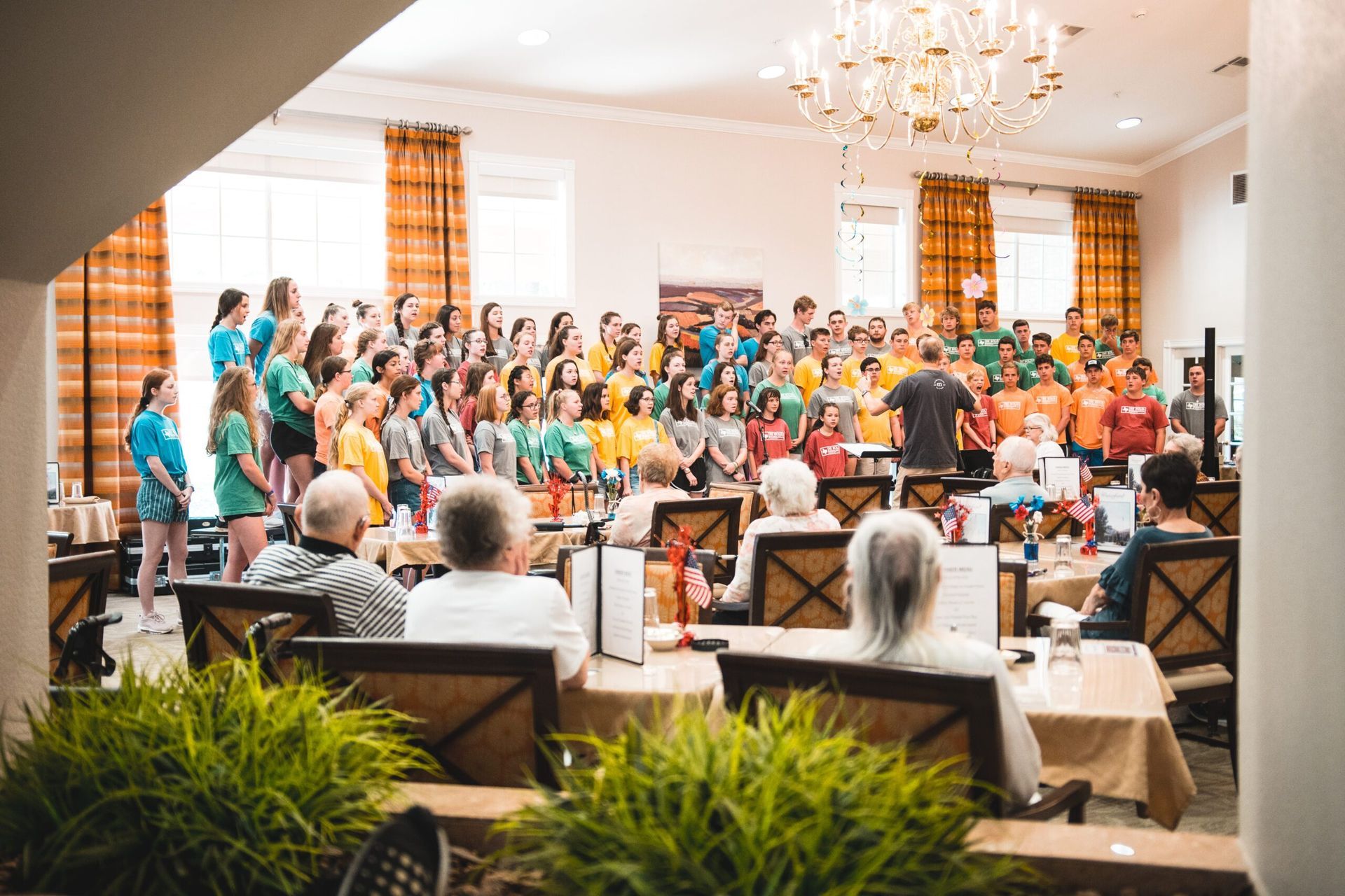 A large group of people are sitting at tables in a room watching a choir perform.