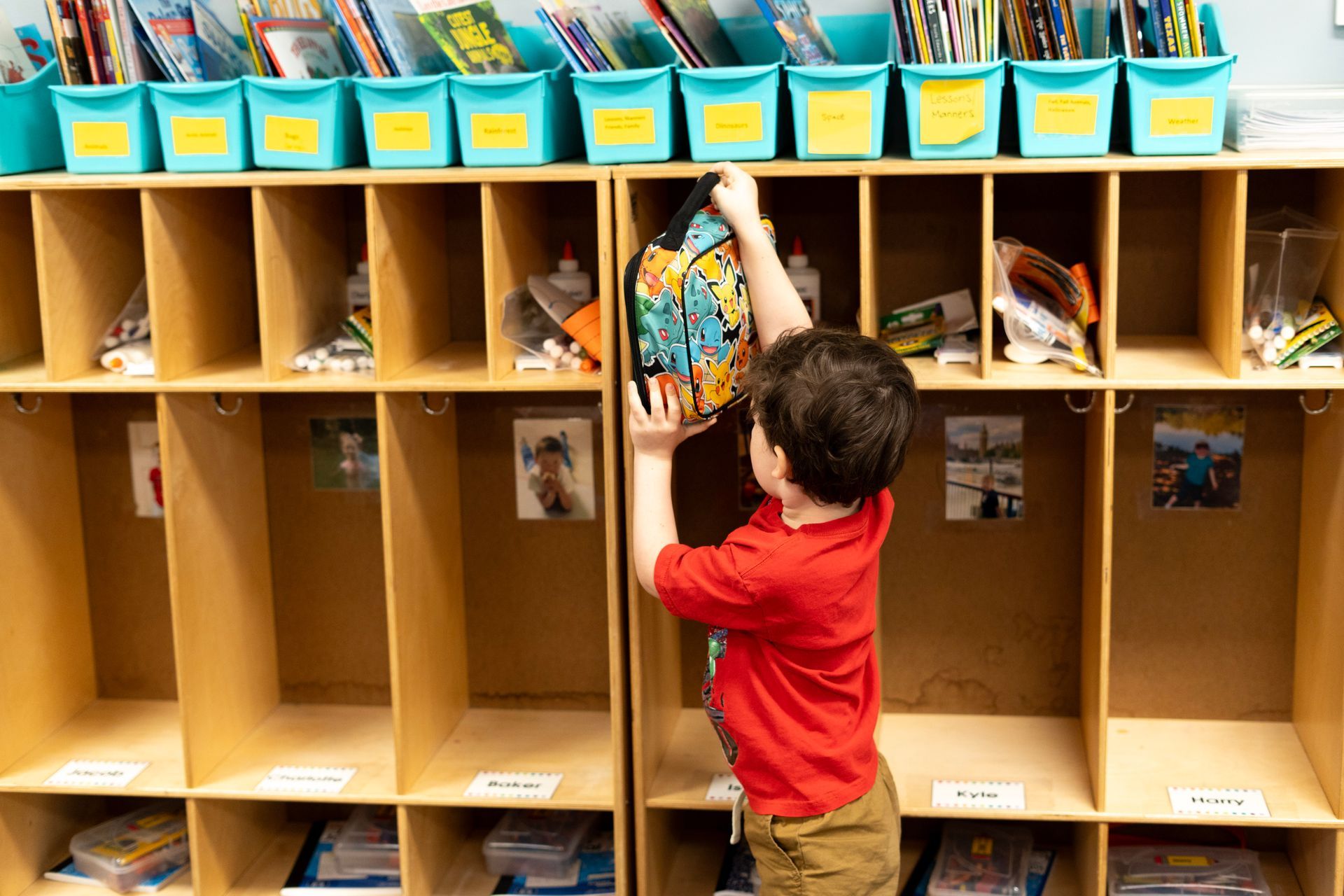 A young boy is putting a backpack in a locker in a classroom.
