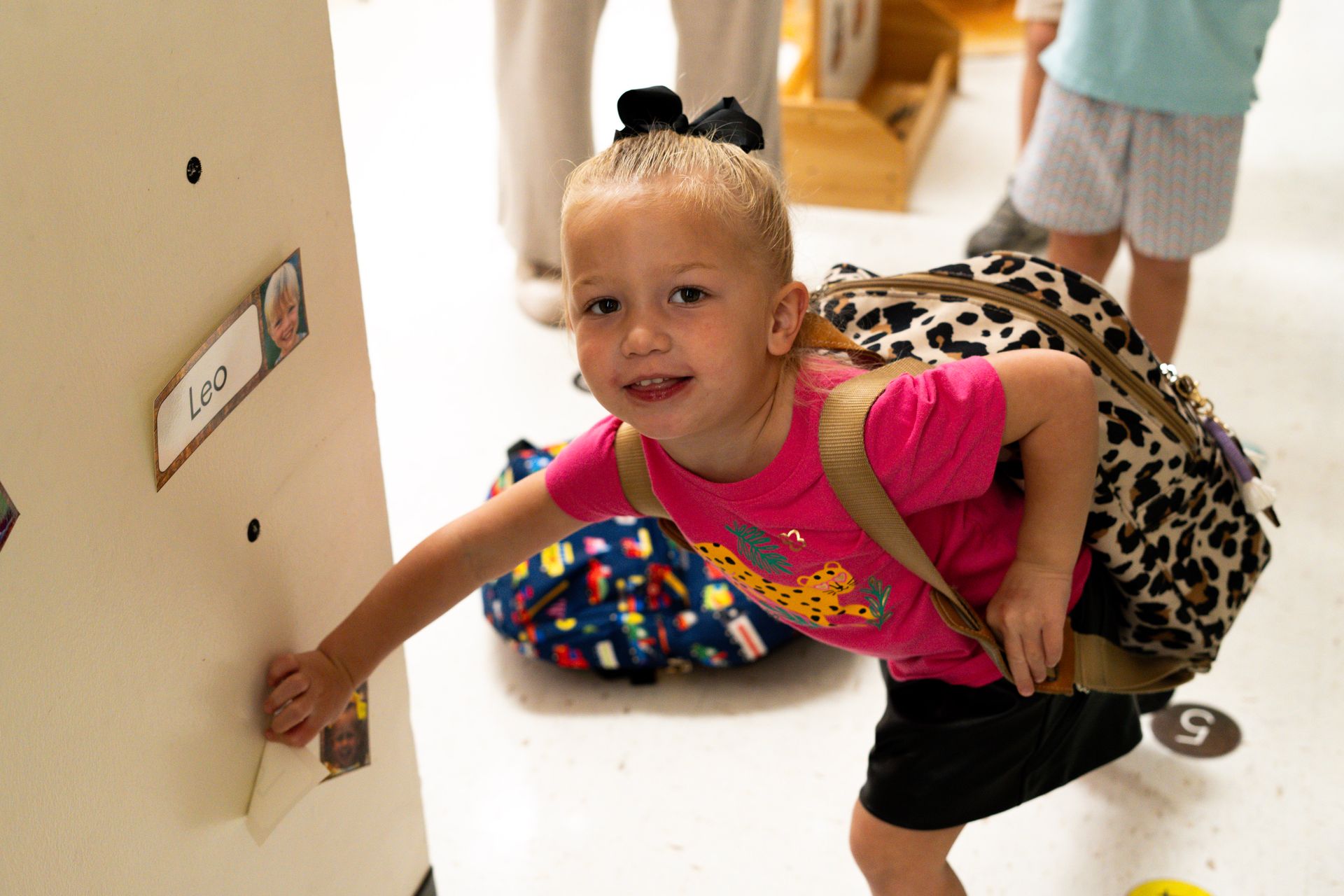 A little girl with a backpack is reaching for a name tag on a door.