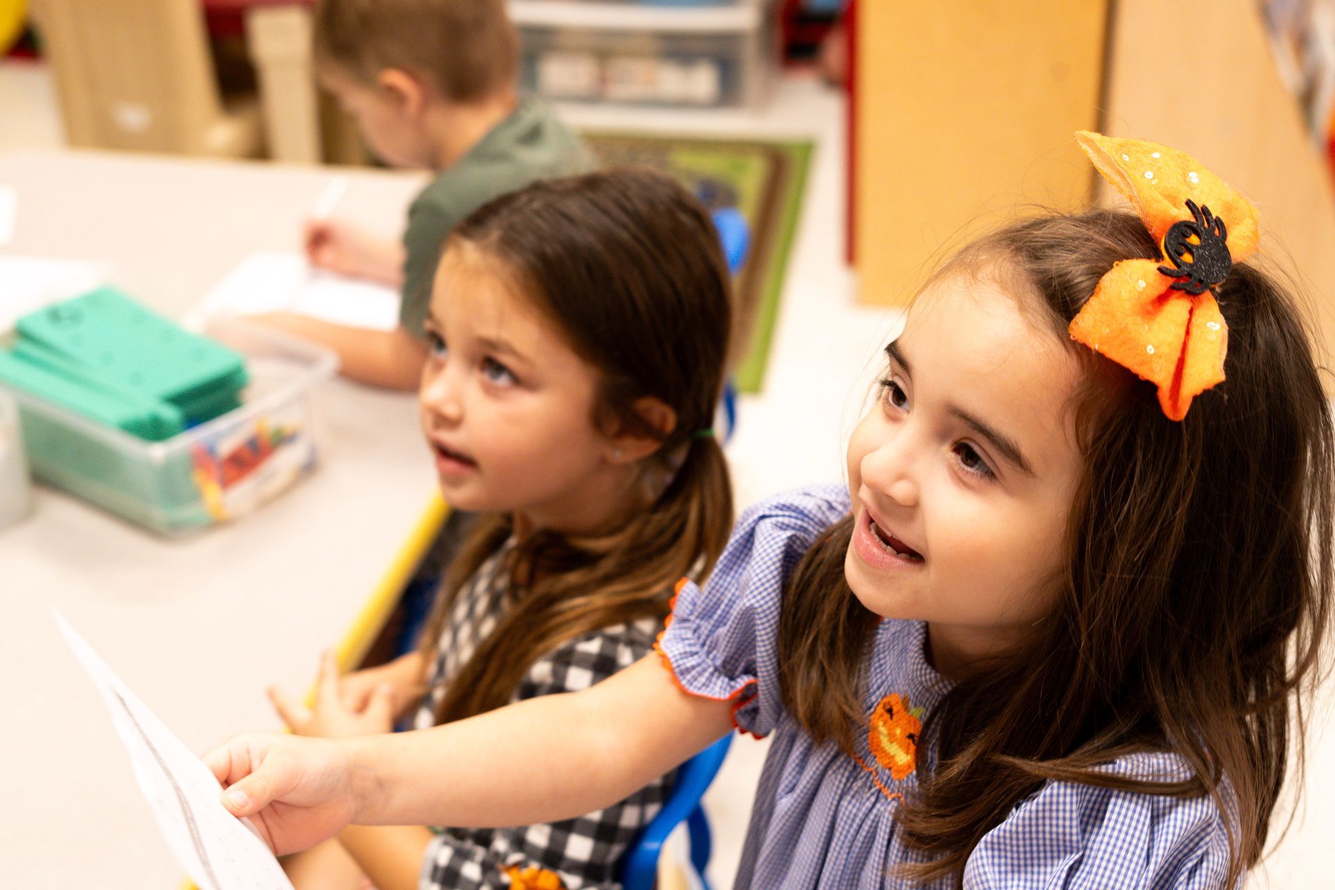 Two little girls are sitting at a table in a classroom looking at a book.