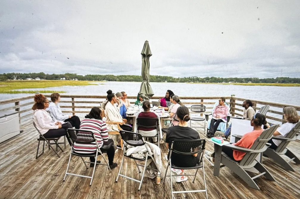 A group of people are sitting around a table on a deck overlooking a body of water.