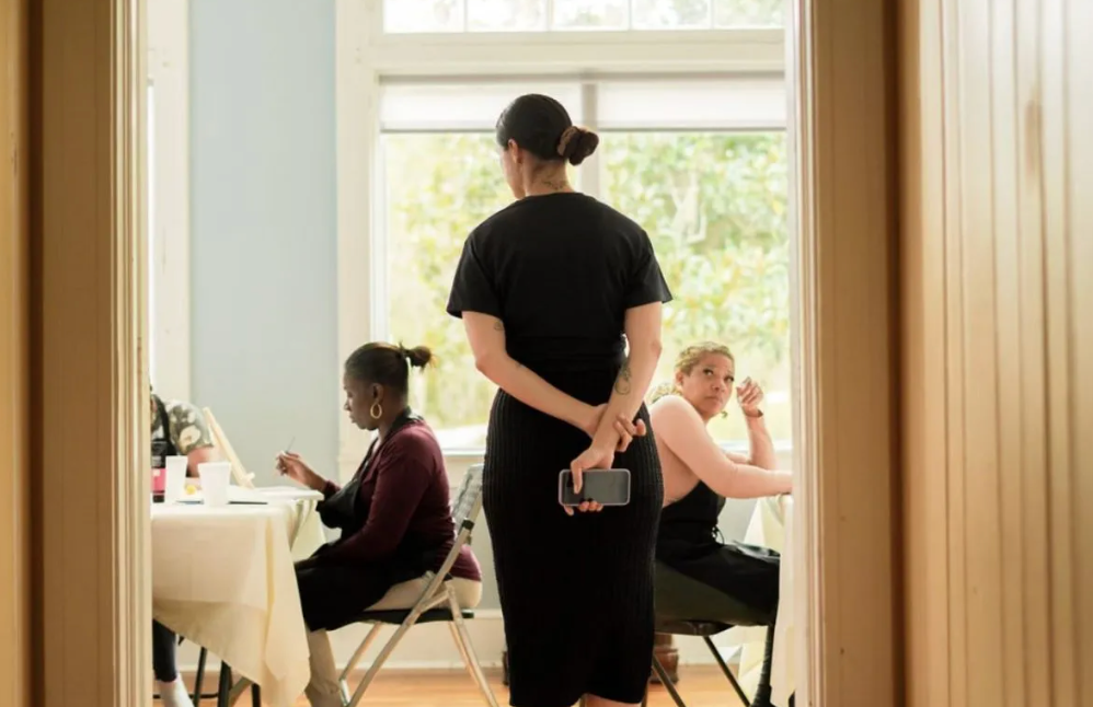 A woman is standing in a room with two women sitting at tables.