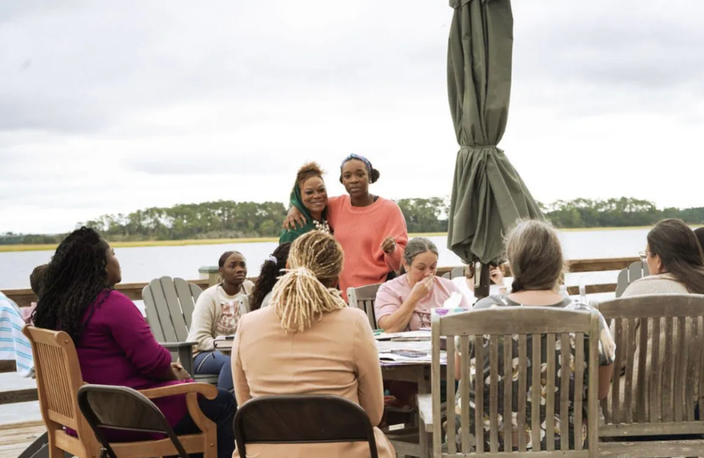 A group of people are sitting around a table in front of a body of water.