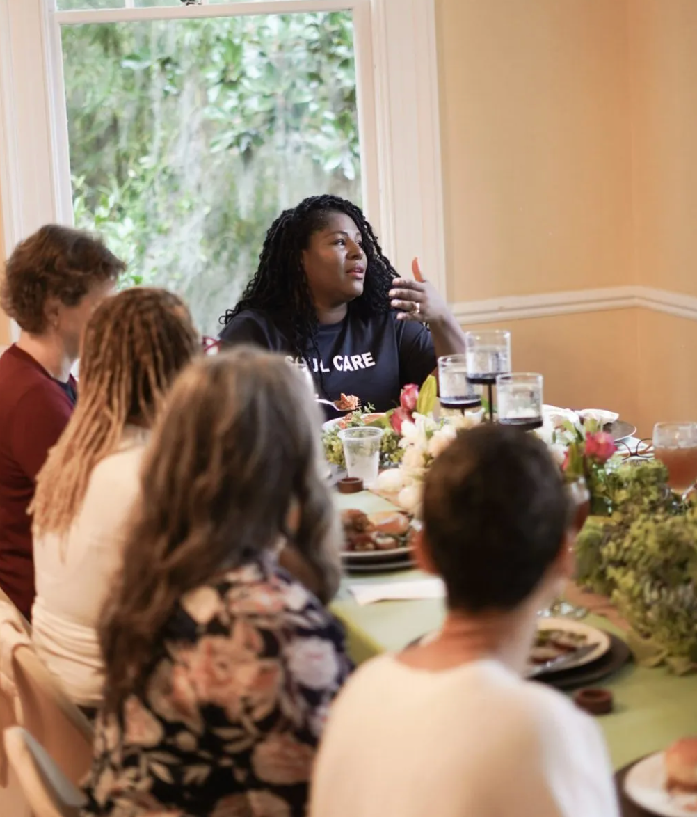 A woman wearing a carol shirt sits at a table with other people