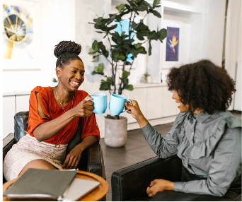 Two women are sitting in chairs holding cups of coffee and laughing.