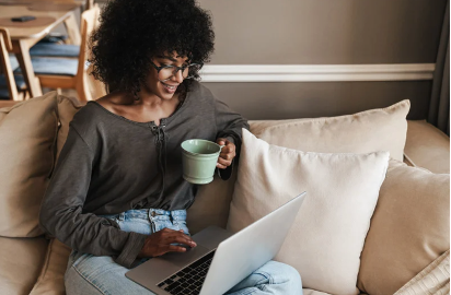 A woman is sitting on a couch using a laptop and drinking coffee.