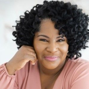 A woman with curly hair is wearing a pink shirt and smiling.