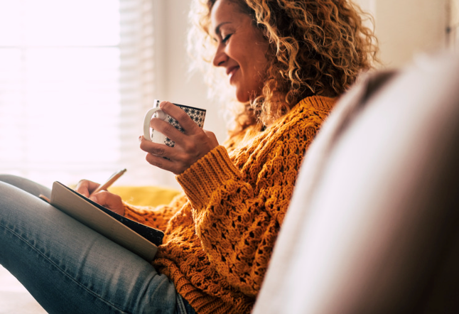 A woman is sitting on a couch holding a cup of coffee and writing in a notebook.