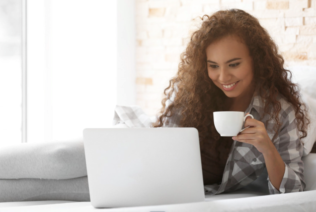 A woman is sitting on a couch drinking coffee and using a laptop computer.