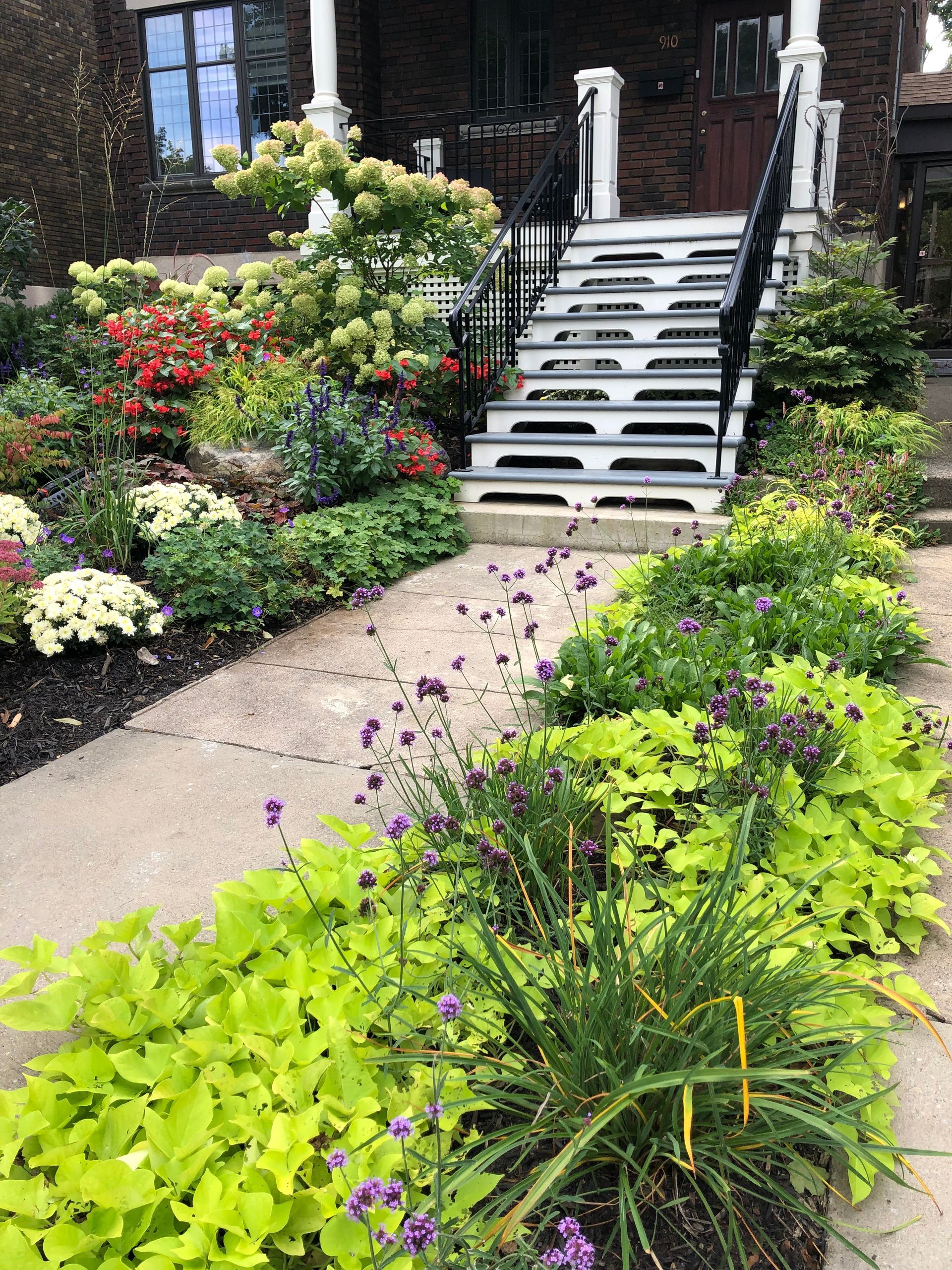 Un jardin avec beaucoup de plantes et de fleurs devant une maison avec des escaliers.