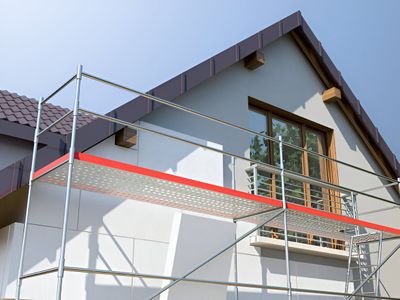 Scaffolding set up against a light gray house with a brown roof; sunlight illuminates the scene.