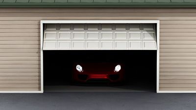 A partially open garage door reveals a dark interior with the headlights of a red car illuminated.