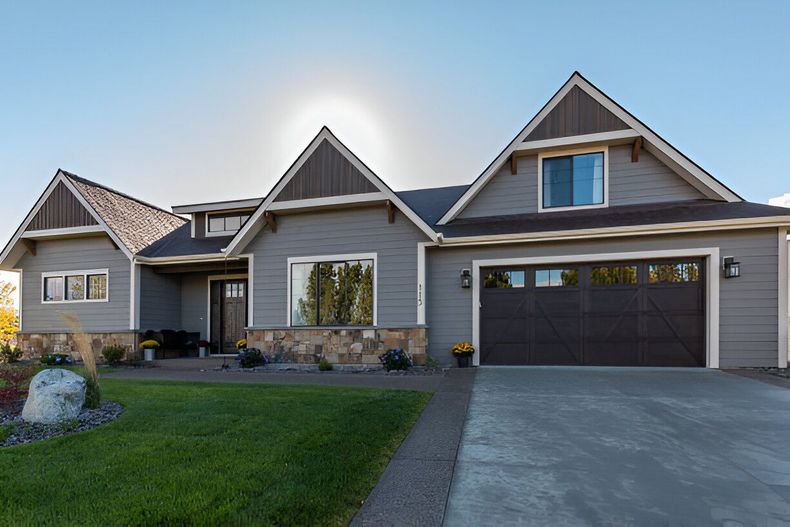 Gray house with a dark garage door and brown roof trim. Features a stone facade, green lawn, and a sunny sky.