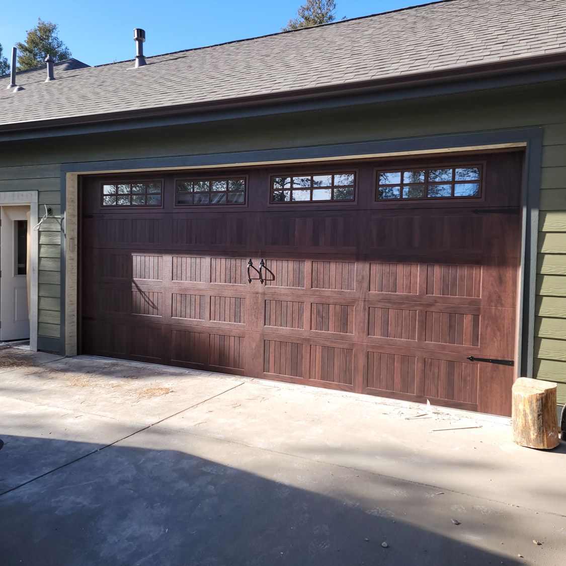 Brown garage door with windows, against a green house. Sunlight. Concrete.