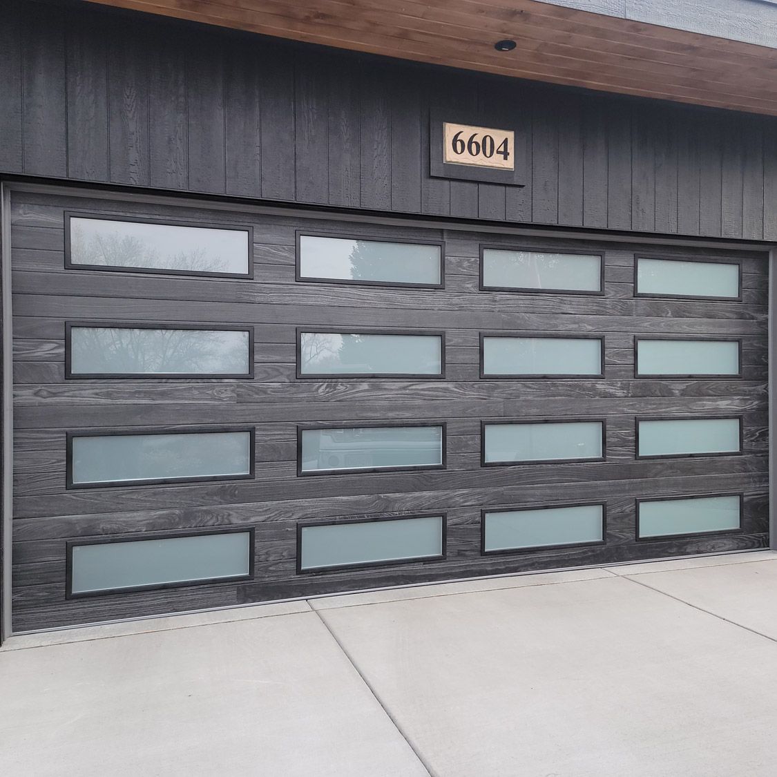Black garage door with rectangular glass windows. Concrete driveway.