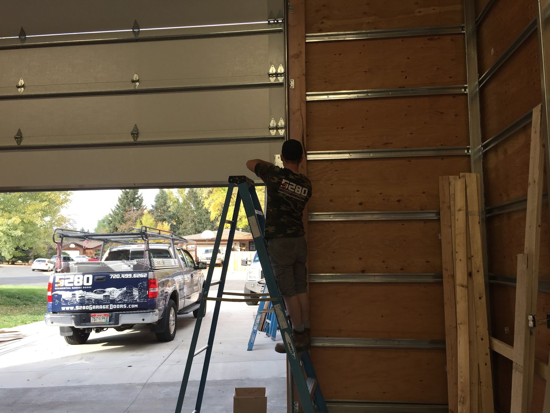 Man on a ladder inside a garage, installing paneling, with a truck parked outside.