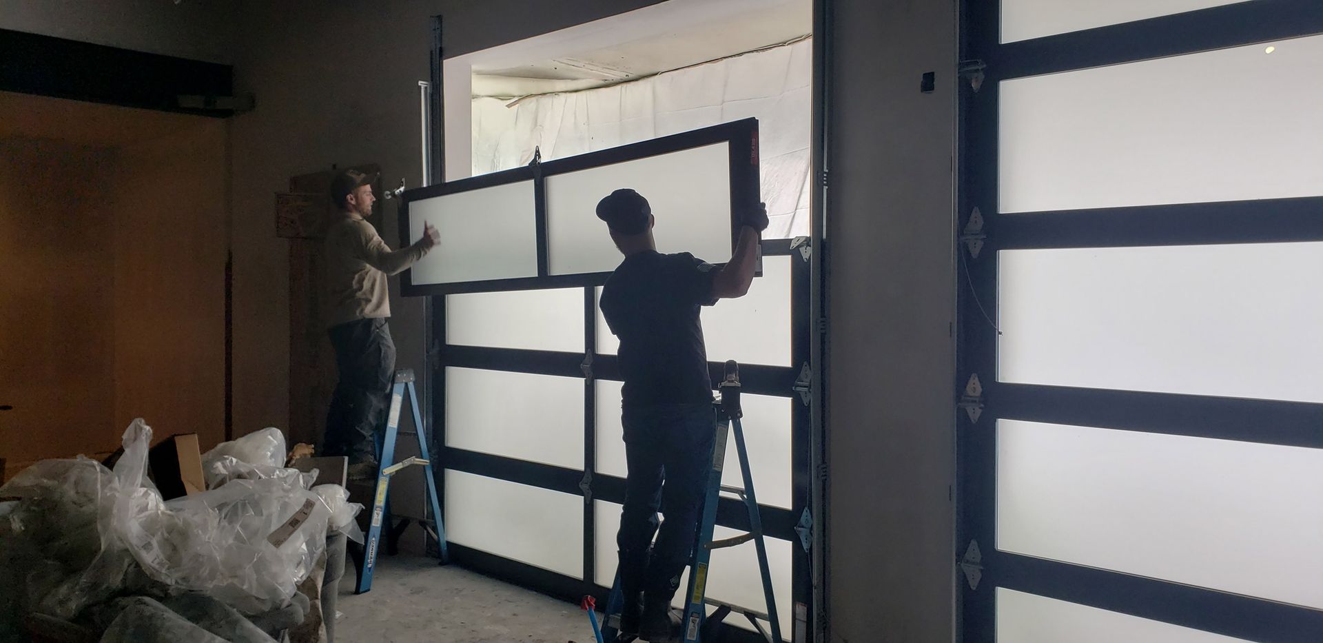 Two men installing a large glass garage door, standing on ladders.