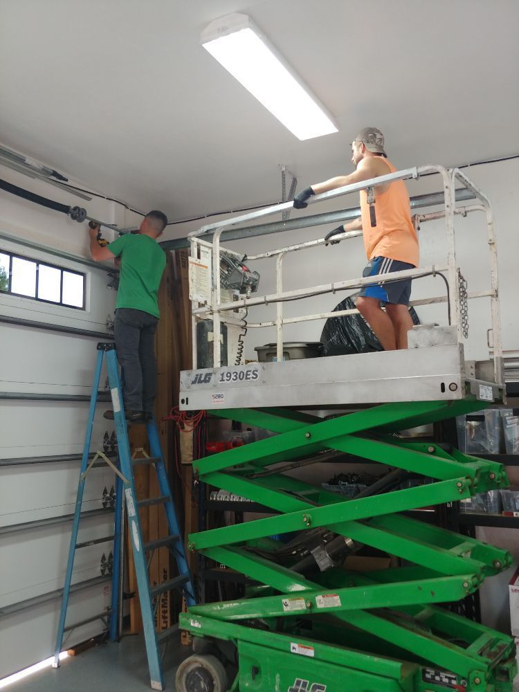 Two workers repair a garage door. One on a ladder, the other on a lift.