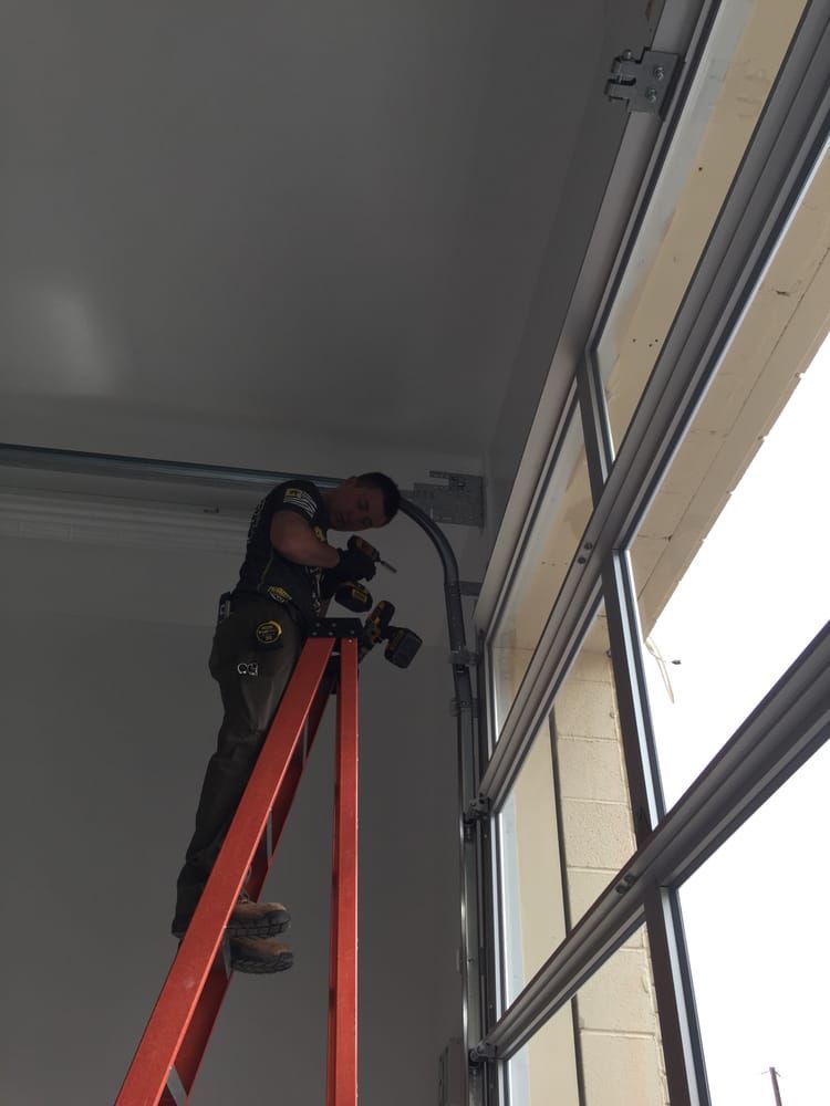 Man on a ladder installing a garage door track next to a glass panel.