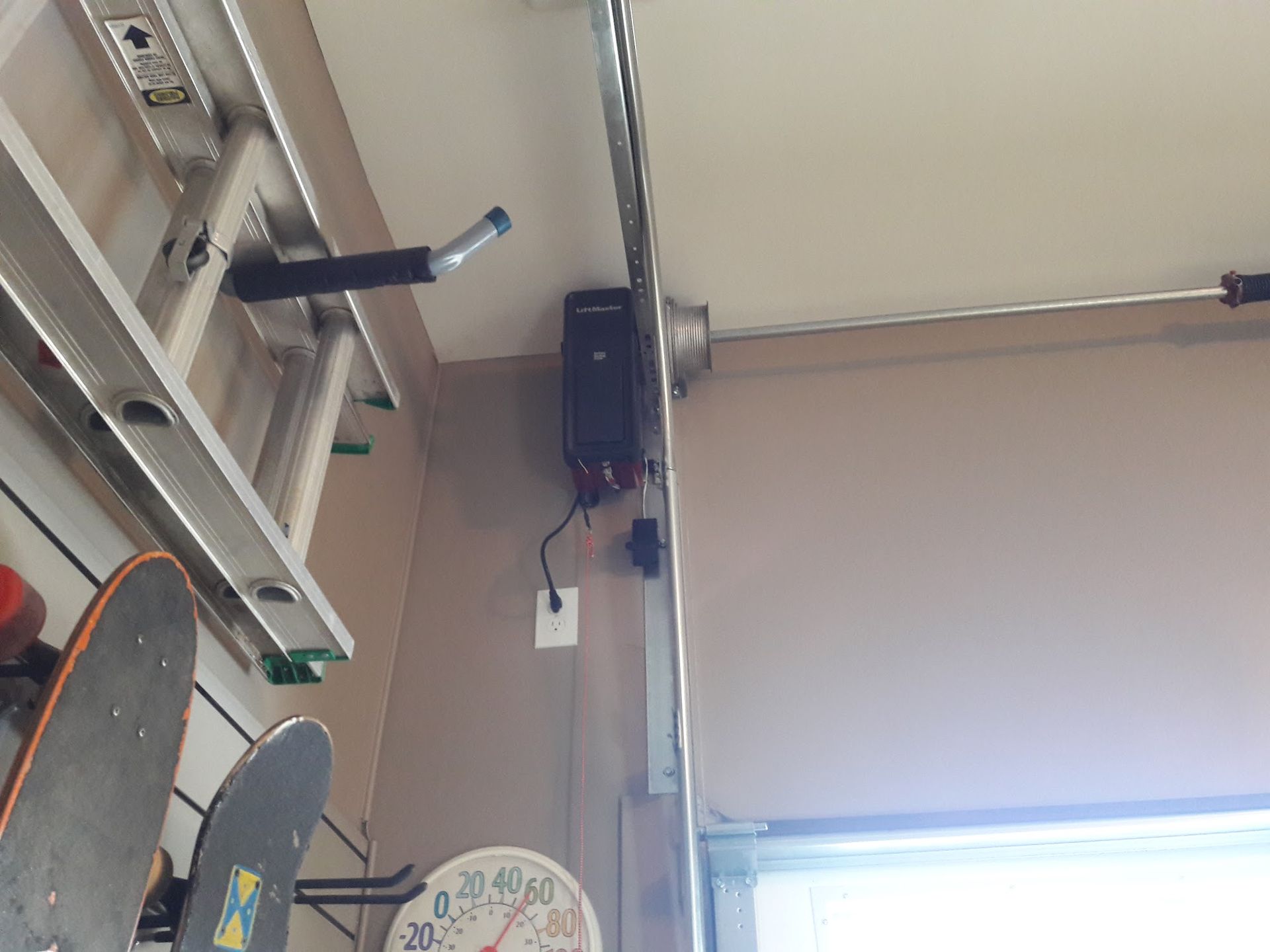 Garage ceiling with a garage door opener, ladder, skateboards, and a thermometer.