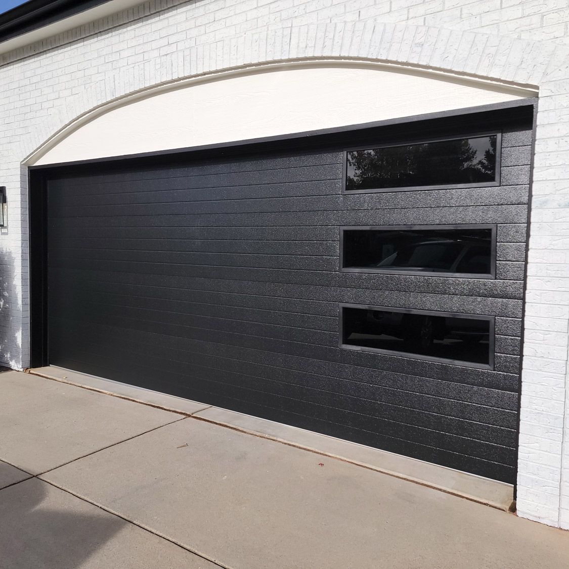 Black garage door with three rectangular windows; white brick exterior.