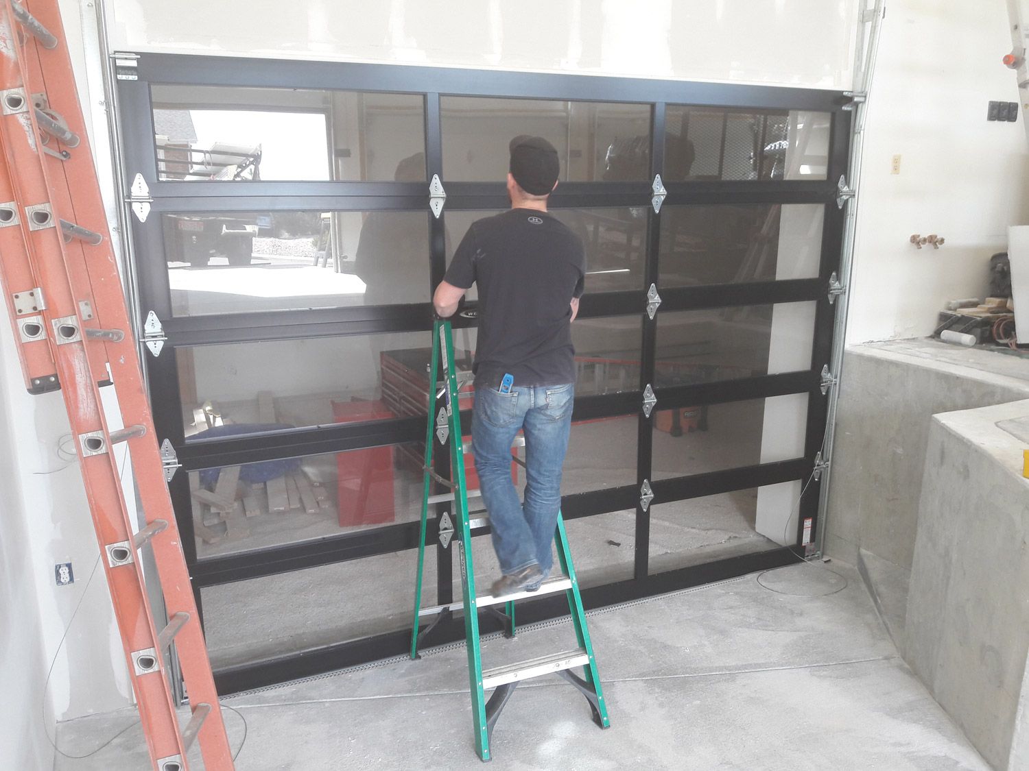 Man on ladder installing a black framed glass garage door. Construction setting, ladder on left.