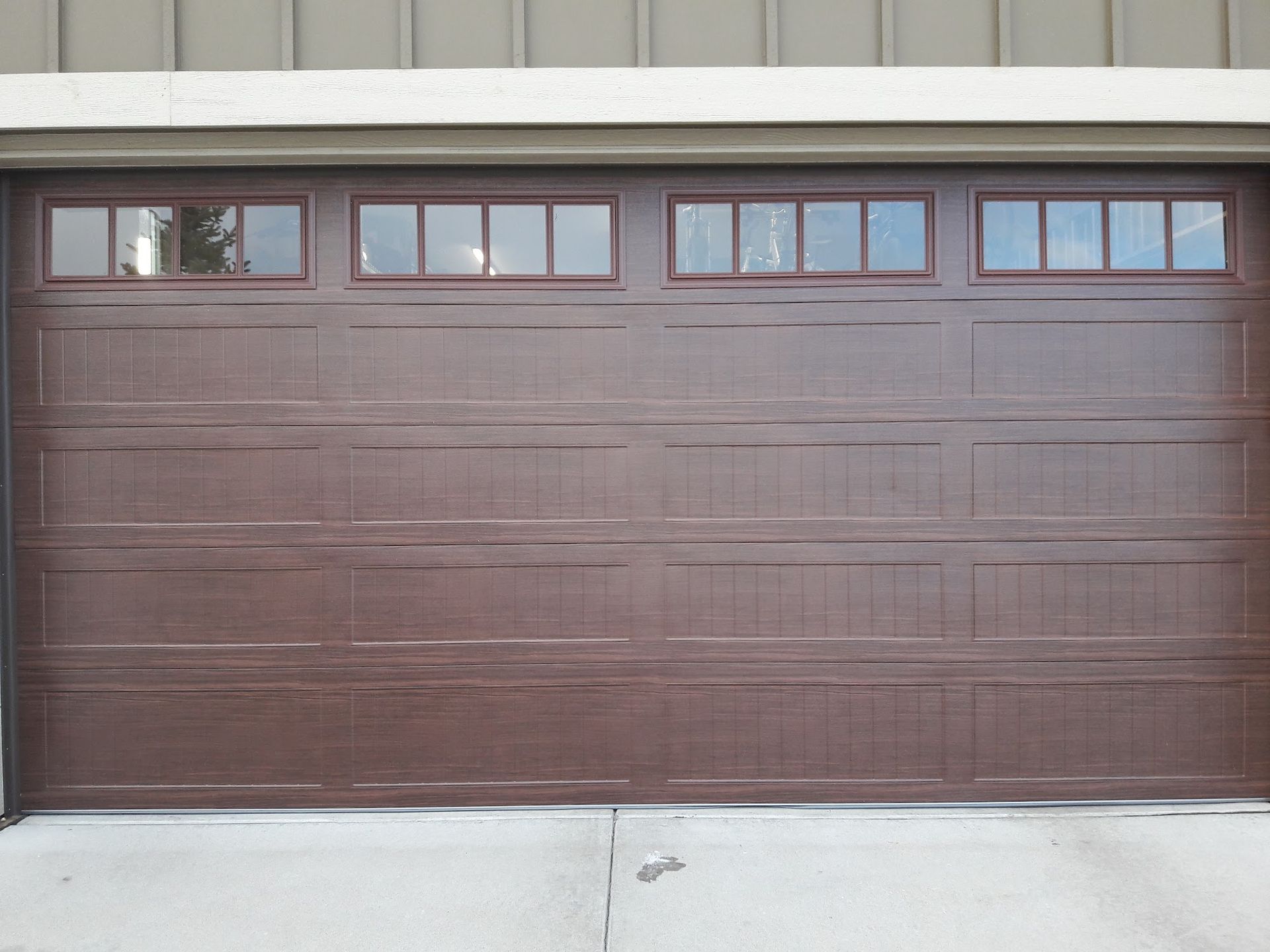 Brown garage door with four top windows. Concrete driveway and light brown exterior siding above.