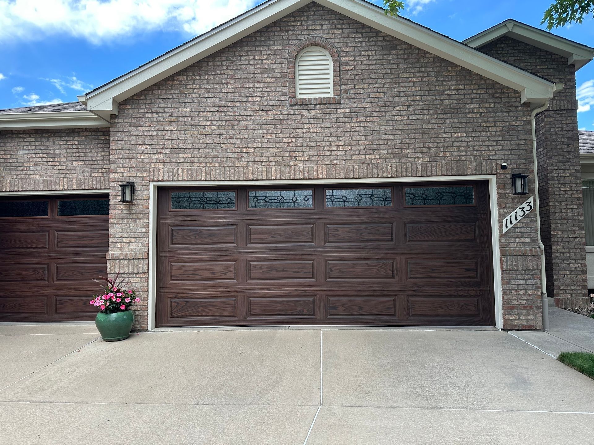 Brown garage doors with decorative glass, set against a brick exterior. Driveway and potted flowers.