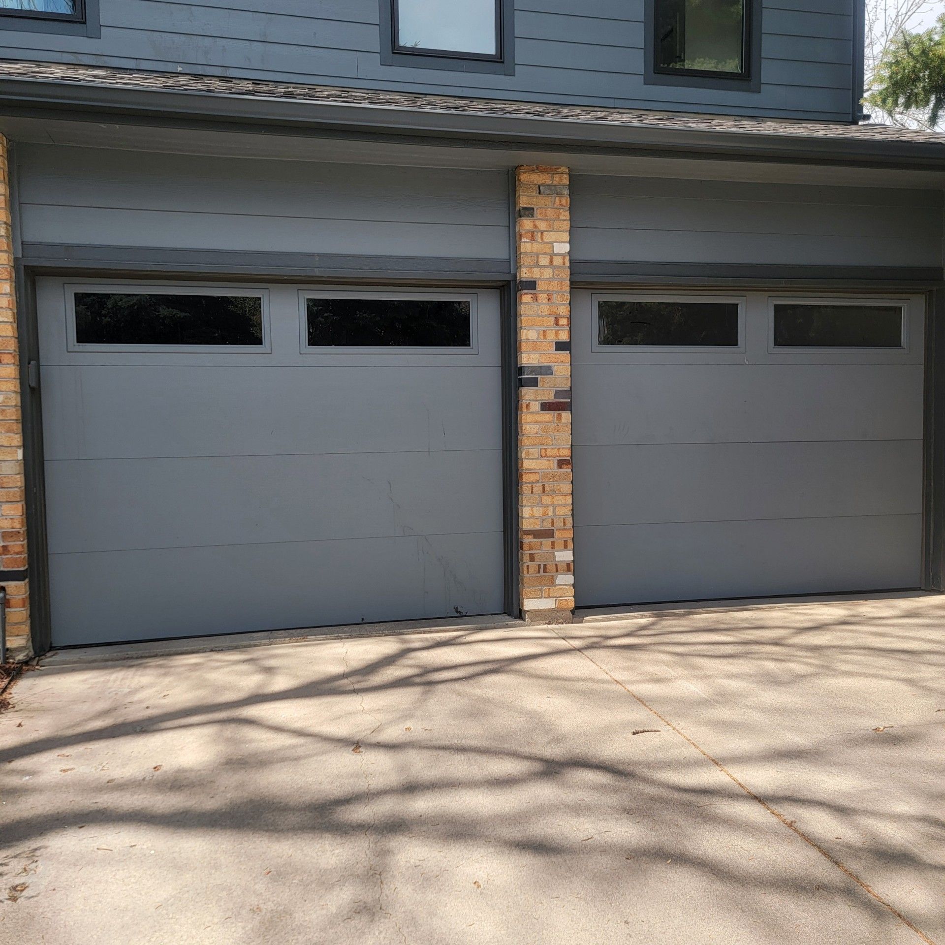 Two gray garage doors with dark window panels, set in a gray house, next to a brick pillar.
