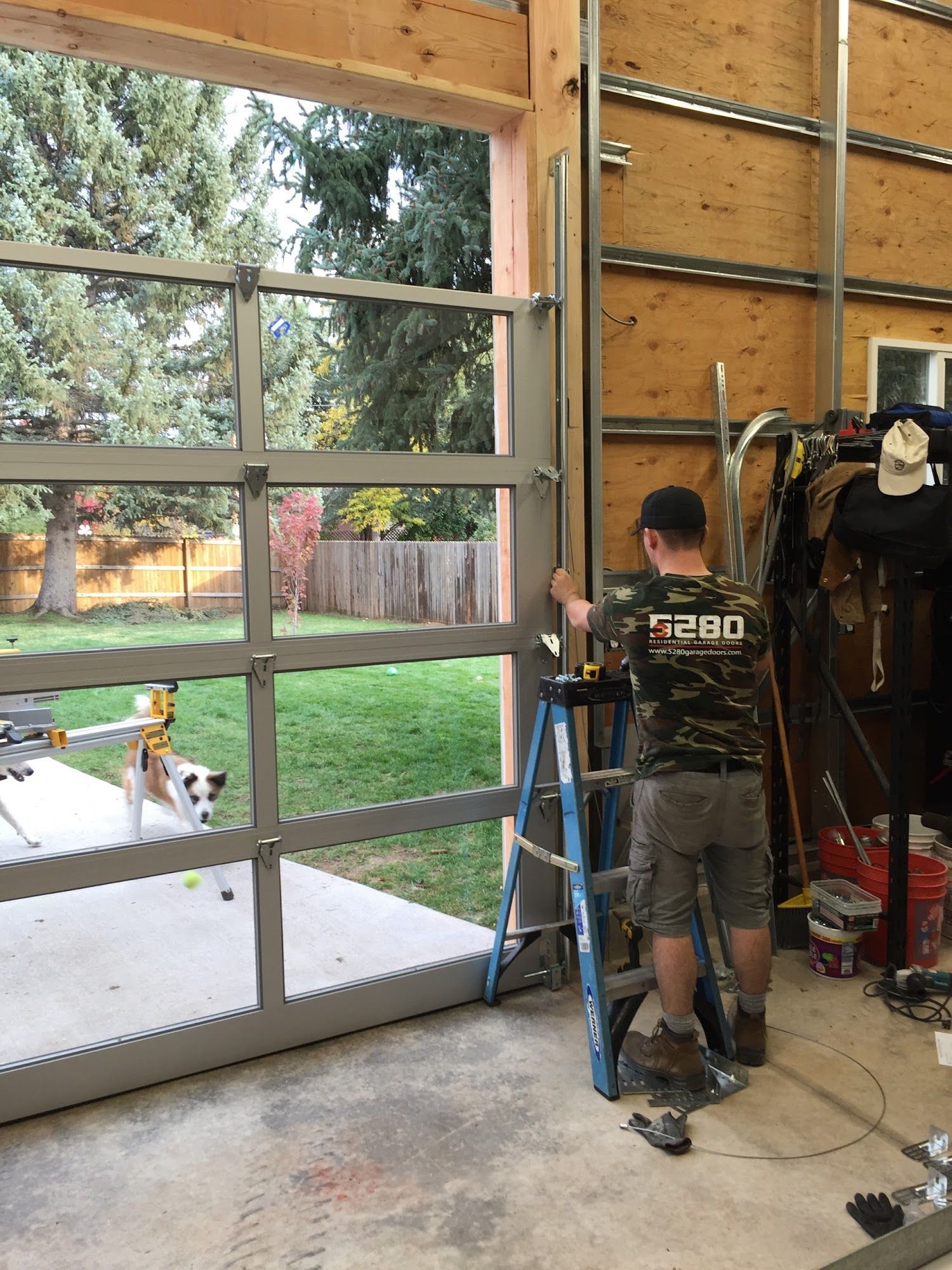 Man installing a glass garage door, standing on a ladder. A dog watches from outside the open door.