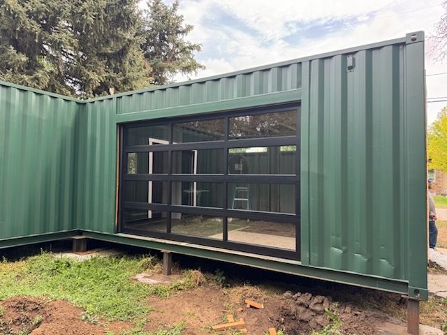 Green shipping container with a large glass door, set on a raised foundation.