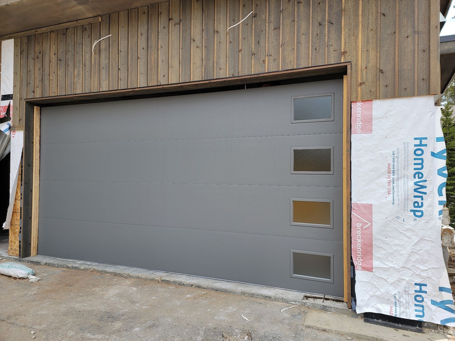 Gray garage door with small square windows, under construction on a wooden-sided building.